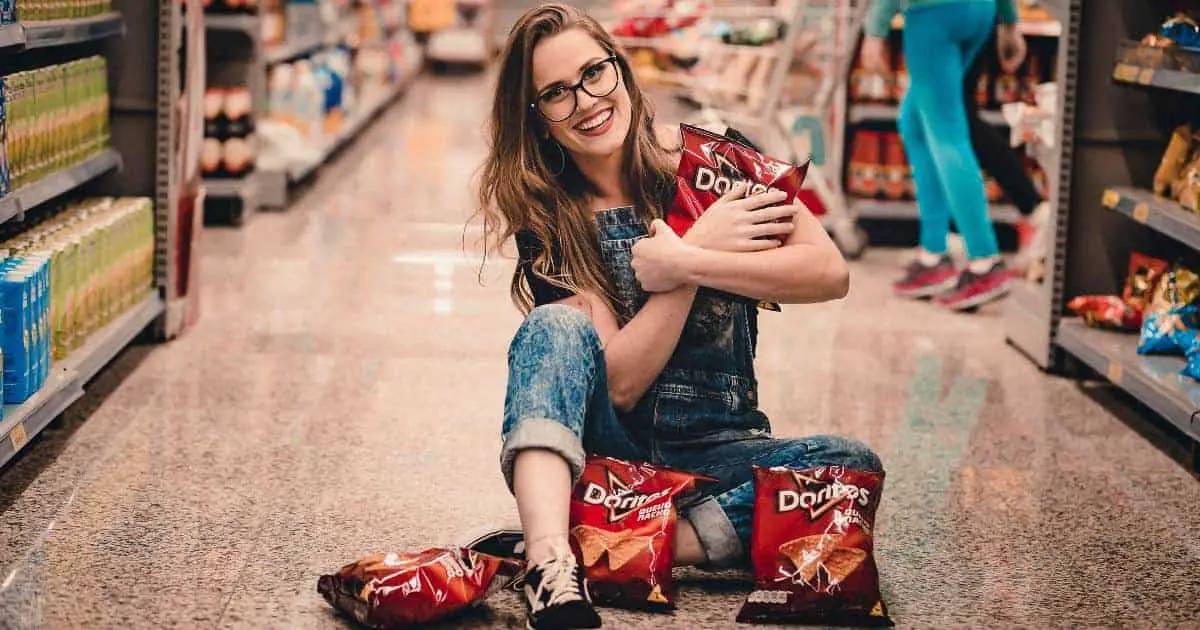 Smiling Woman Sitting in the Middle of Shopping Aisle Holding Dorito Chips